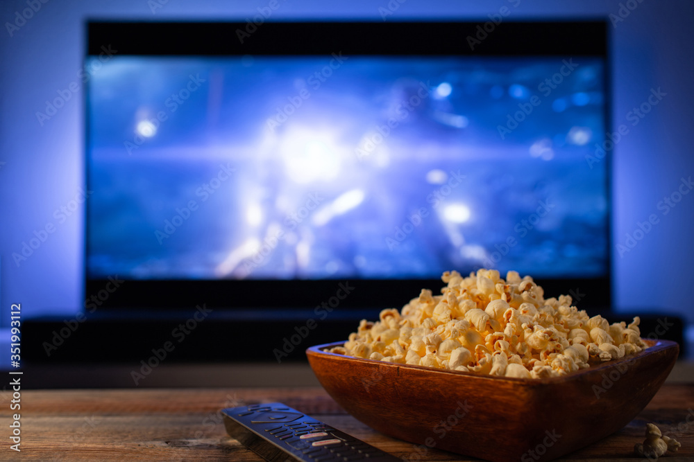 A wooden bowl of popcorn and remote control in the background the TV works. Evening cozy watching a movie or TV series at home A wooden bowl of popcorn and remote control in the background the TV works. Evening cozy watching a movie or TV series at home