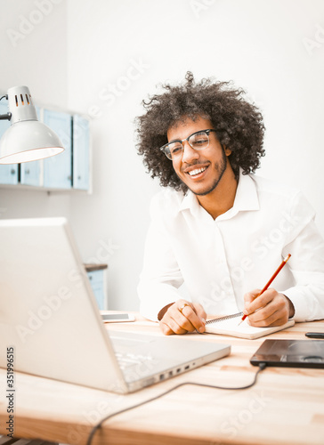 Copywriter takes notes in a notebook while sitting at a desk with a laptop computer on it. Young author man working on new project. Work from home concept. Toned image