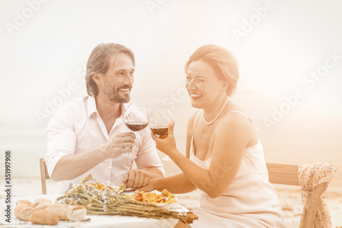 Happy man and woman drink red wine at seaside cafe outdoors. Smiling middle aged couple have date sitting holding hands near the sea. Toned image