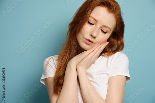 A red-haired girl in a white T-shirt stands on a blue background, closing eyes, bowing head on side and pinning palms to cheek