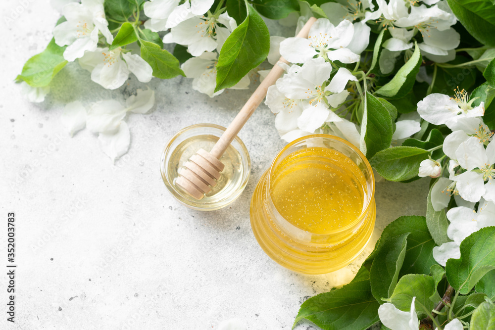 Flower honey in a glass jar on the background of flowering branches. Liquid honey in a glass jar on a light gray table. Jar of honey close-up. Top view with space for text