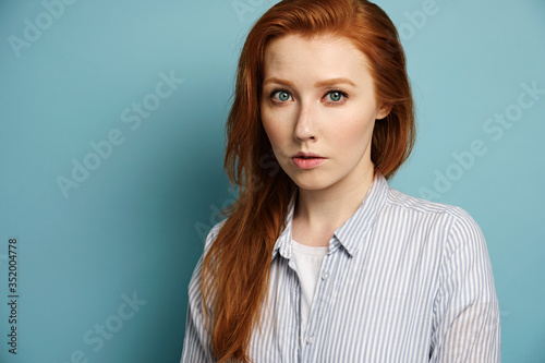 The red-haired girl in the shirt stands in a half-turn on a blue background and looks into the frame.