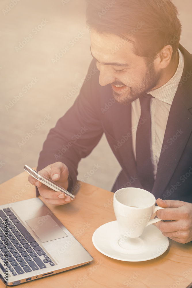 Happy businessman reading messages on mobile phone. Laughing man drinks coffee in street cafe and chatting on smartphone while working on computer. Freelance concept. Toned image Happy businessman reading messages on mobile phone. Laughing man drinks coffee in street cafe and chatting on smartphone while working on computer. Freelance concept. Toned image