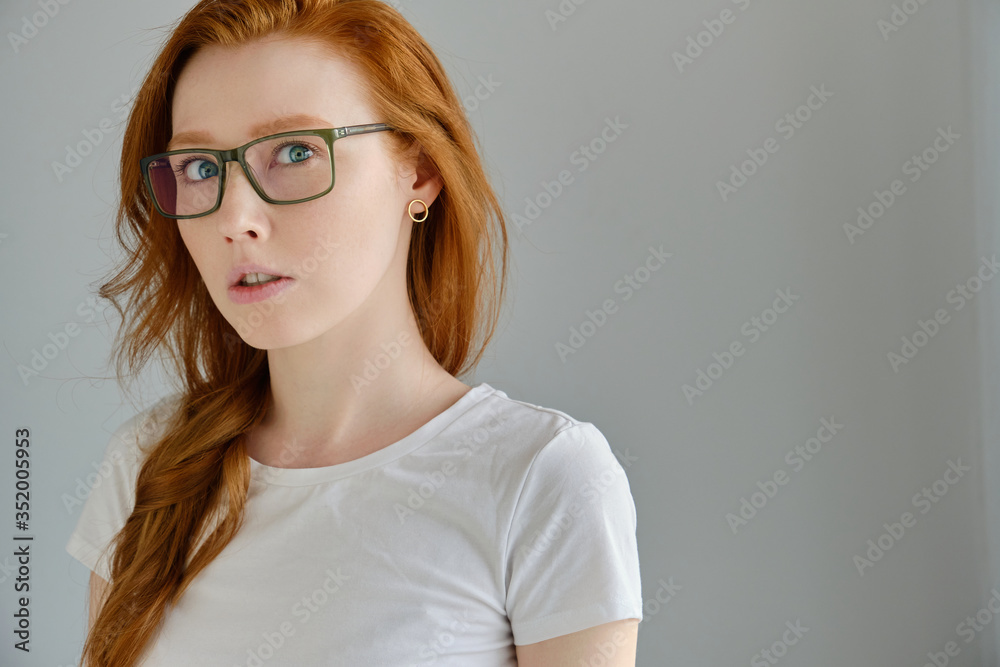 A red-haired girl in glasses and a white T-shirt stands on a gray background in a half-turn and looks surprised into the frame A red-haired girl in glasses and a white T-shirt stands on a gray background in a half-turn and looks surprised into the frame