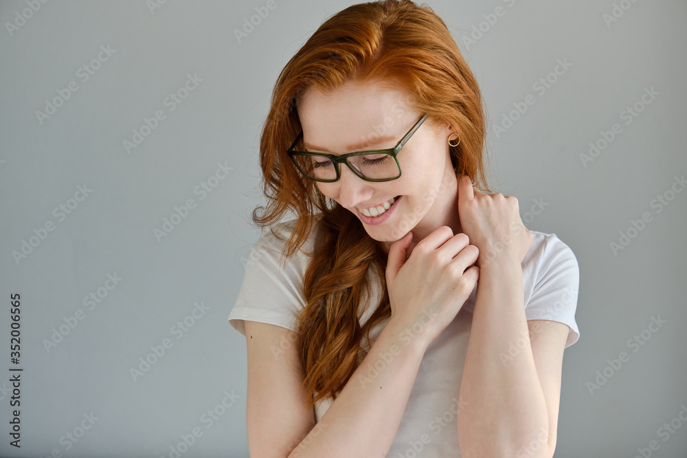A red-haired girl in a white T-shirt and glasses stands on a gray background and laughs with her head down. A red-haired girl in a white T-shirt and glasses stands on a gray background and laughs with her head down.