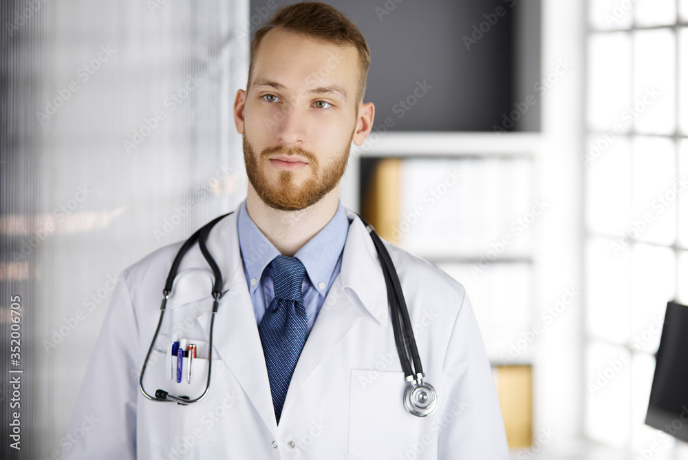 Red-bearded doctor standing straight in clinic near his working place. Portrait of physician. Medicine and healthcare concept Red-bearded doctor standing straight in clinic near his working place. Portrait of physician. Medicine and healthcare concept