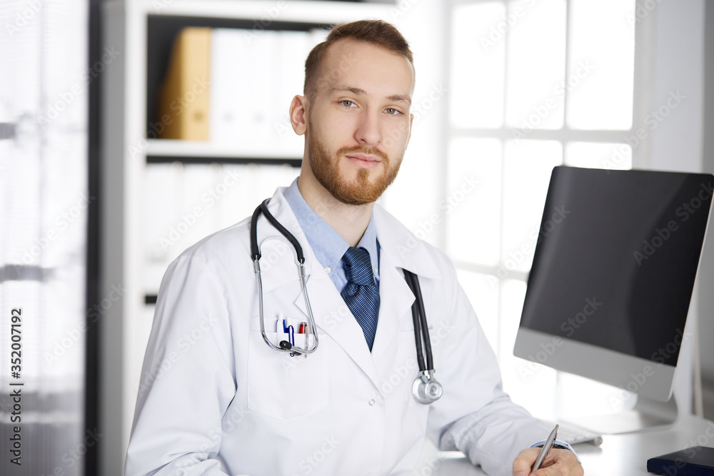 Friendly red-bearded doctor sitting and writing at clipboard in clinic. Perfect medical service in hospital. Medicine concept Friendly red-bearded doctor sitting and writing at clipboard in clinic. Perfect medical service in hospital. Medicine concept