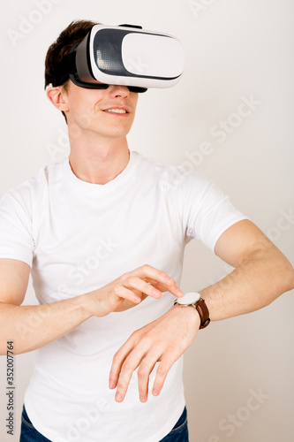 Young man developer using virtual reality headset interacts with augmented things orienting in three dimensional space while sitting on his desk with keyboard and laptop on white background.