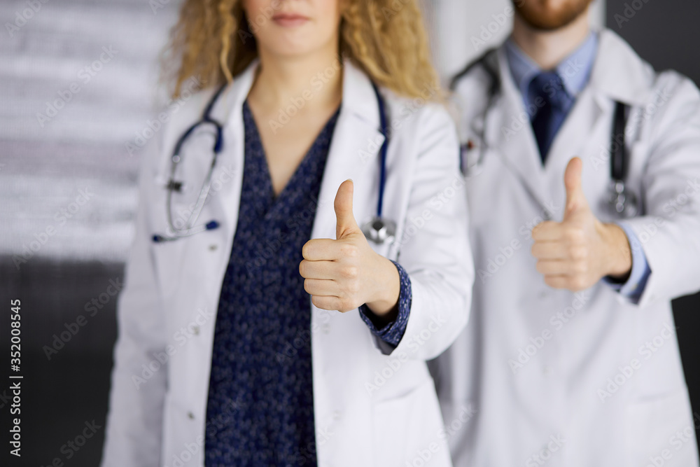 Two doctors standing with thumbs up in hospital office. Medical help, countering viral infection and medicine concept