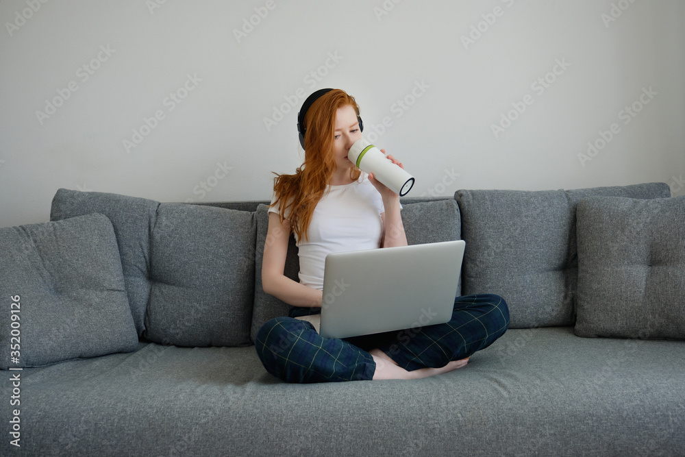 A red-haired girl with headphones sits at home on the couch in pajamas with a laptop and drinks from a thermo cup A red-haired girl with headphones sits at home on the couch in pajamas with a laptop and drinks from a thermo cup