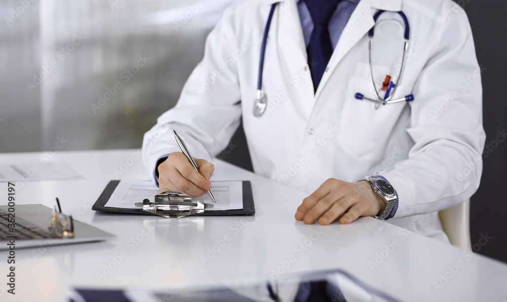 Unknown male doctor sitting and working with clipboard of medication history record in clinic at his working place, close-up. Young physician at work. Perfect medical service, medicine concept Unknown male doctor sitting and working with clipboard of medication history record in clinic at his working place, close-up. Young physician at work. Perfect medical service, medicine concept
