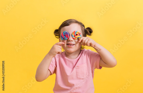 A laughing girl with ponytails closes her eyes with lollipops on a yellow background