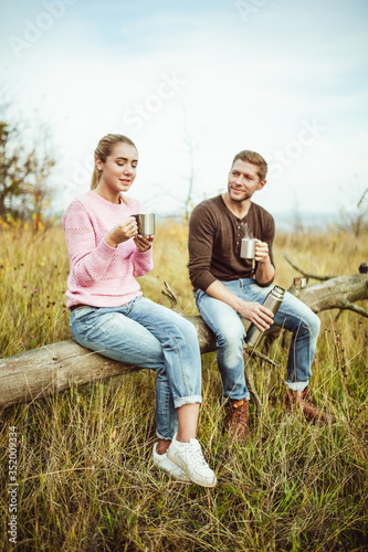 Tea party or coffee drinking outdoors. Cheerful couple drink hot coffee or tea communicate sitting on a wooden log outdoors