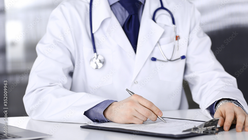 Unknown male doctor sitting and working with clipboard in clinic at his working place, close-up. Young physician at work. Perfect medical service, medicine concept Unknown male doctor sitting and working with clipboard in clinic at his working place, close-up. Young physician at work. Perfect medical service, medicine concept
