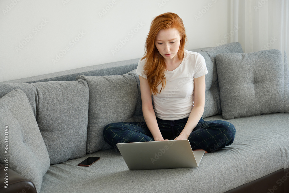 A red-haired girl sits cross-legged in sofa in home clothes and looks into a laptop. A red-haired girl sits cross-legged in sofa in home clothes and looks into a laptop.