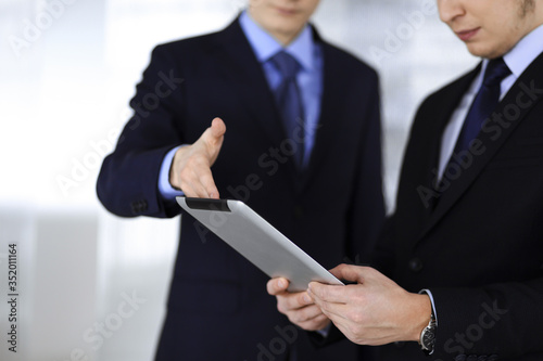 Business people use a tablet computer for discussion of their new project, standing in a modern office. Unknown businessman or male entrepreneur with a colleague at workplace. Teamwork and partnership