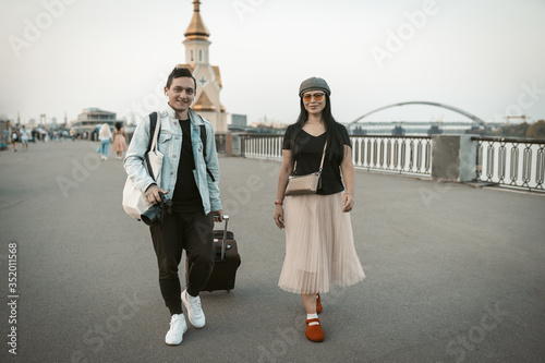 Two travelers are walking along the promenade of a european city. Smiling man and woman with backpacks and suitcases go along the riverbank looking at camera. Adventure concept