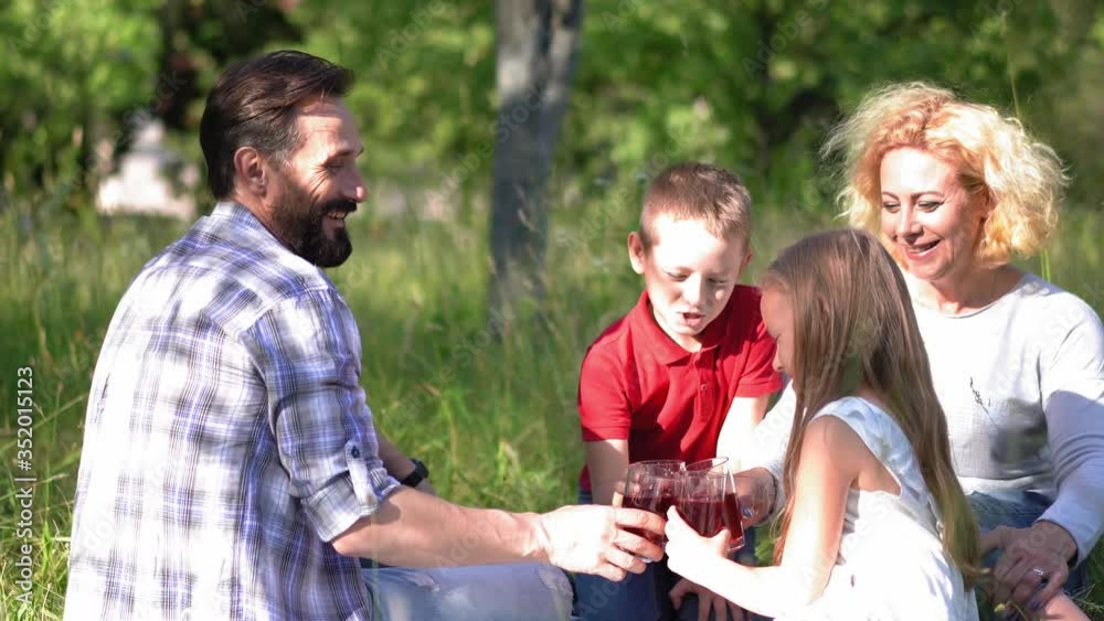 Family on a picnic with mother in a wheelchair spends time with two children boy and girl drinking juice on the lawn in a nature park. Family concept.