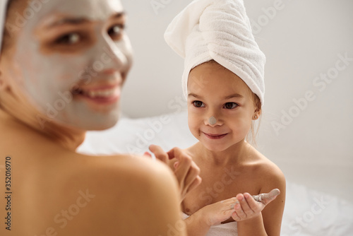 A young caucasian mother and little daughter with wrapped hair in white bath towels apply a clay mask on the mother faces