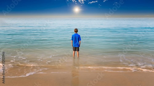 Young boy staring our towards to the horizon with his feet in the surf and a beautiful blue sea and a dramatic sky with a sun star in the distance.