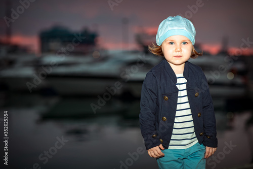 a little beautiful girl, in sea-style clothes, walks in the summer at sunset, on the beach near the yachts 