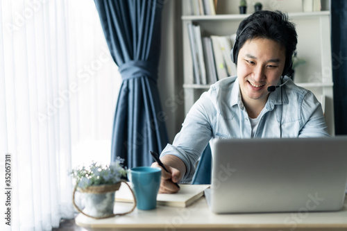 Asian businessman talking to colleague team in video call conference writing note on book with smile face. Man using computer laptop and headphone for online meeting. Smart working from home concept.
