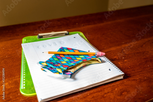 Face mask resting on top of back to school supplies of paper, pencil and a clip board photo
