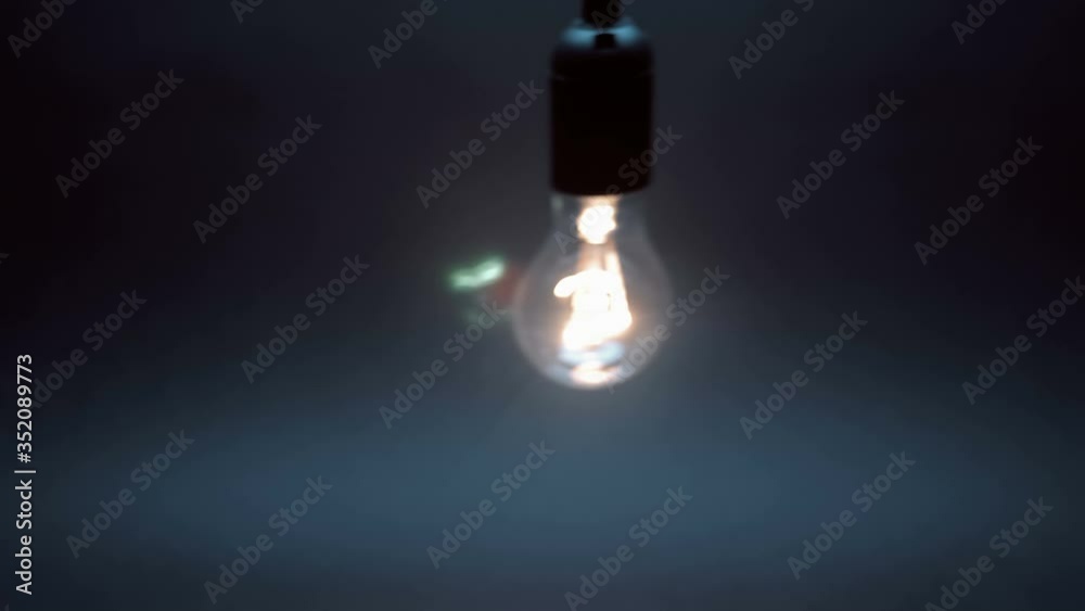 Classic glass lamp with tungsten filament lights up, swaying on the wire on a black background. Closeup