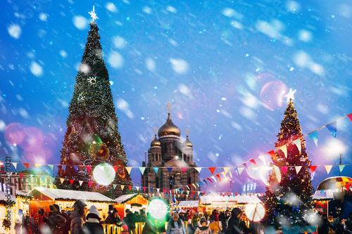 square with two Christmas trees and a church in the middle, in the foreground people, lights, it is snowing © Anna Rudoy