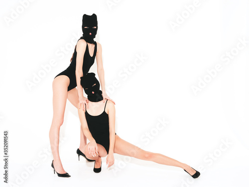 Two beautiful sexy women in black swimwear bathing suit. Models wearing bandit balaclava mask.Hot girls posing near white wall in studio.Seductive female in nice lingerie.Crime and violence