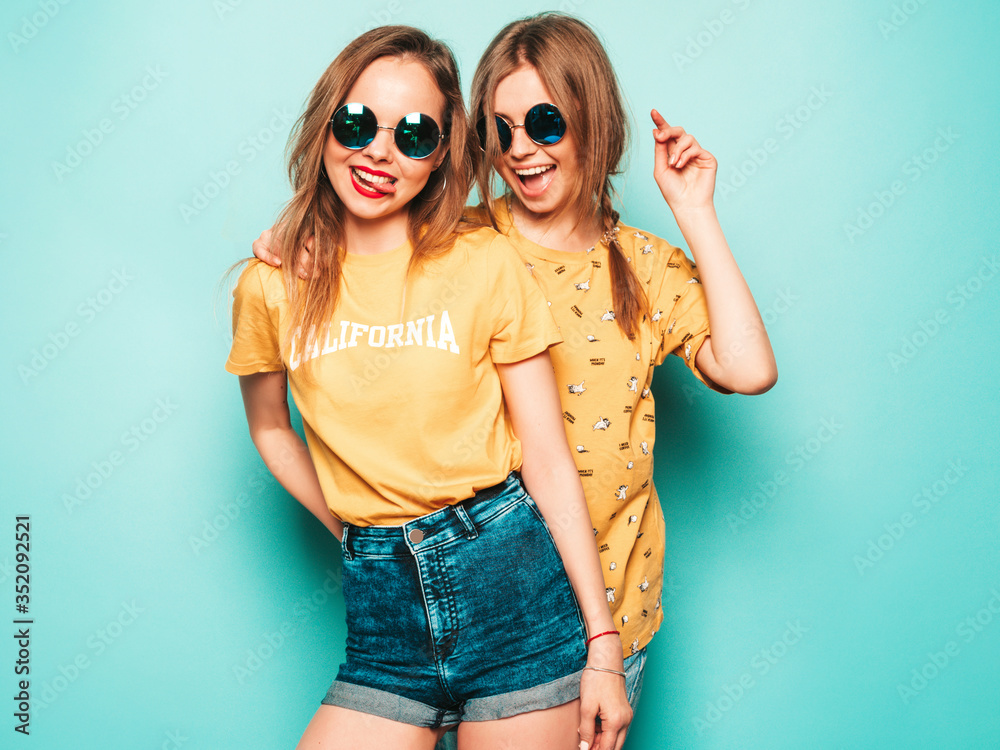 Two young beautiful smiling hipster girls in trendy summer yellow T-shirts. Sexy carefree women posing near blue wall. Trendy and positive models having fun in sunglasses