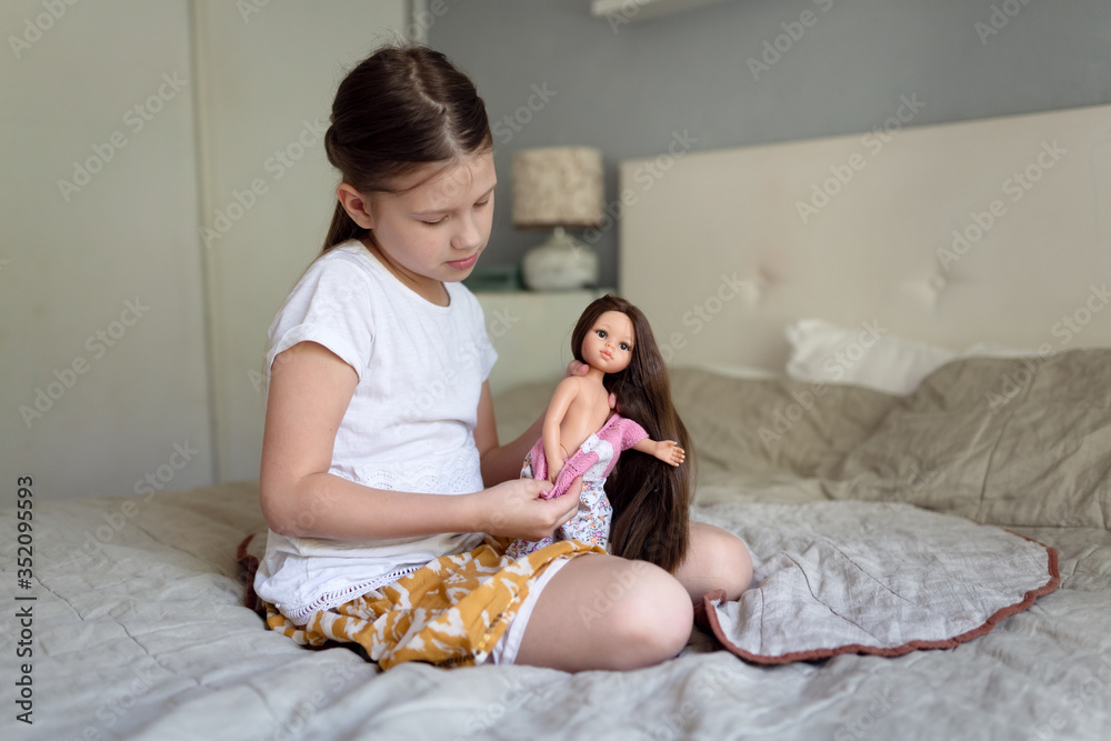 Caucasian girl dresses up doll with long hair, girl plays with dolls on bed in bedroom. Traditional kids games for girls Caucasian girl dresses up doll with long hair, girl plays with dolls on bed in bedroom. Traditional kids games for girls