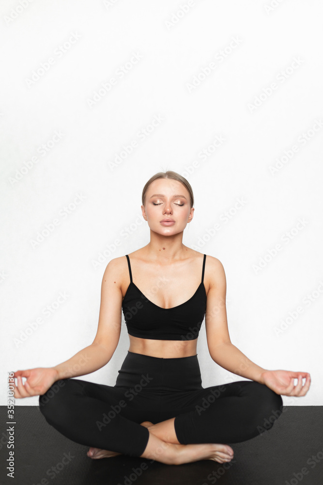 A beautiful caucasian woman with a great figure is meditating on a black yoga mat. White background. Healthy lifestyle and a beautiful female figure. Sportswear.