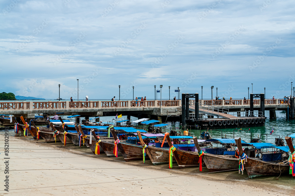 Parking of tieded traditional thai longtail boats with colorful tape, Thailand. Waiting for passenger. Tourists on the pier on blue sky background. Ships, yachts and fishing boats at low tide.