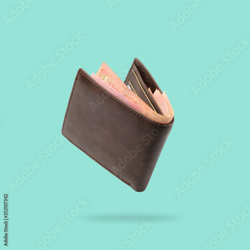 Flying in air Brown genuine leather wallet with banknotes and credit card inside isolated on turquoise background.