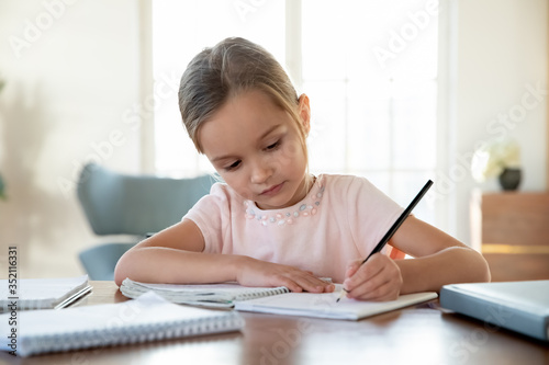 Smart small schoolgirl sit at desk at home studying handwriting in notebook, hardworking focused little girl child write doing homework lessons alone, feel motivated at learning, education concept
