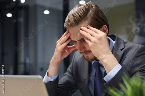 Image of tired young business man in suit keeping face in hands while sitting in the office.