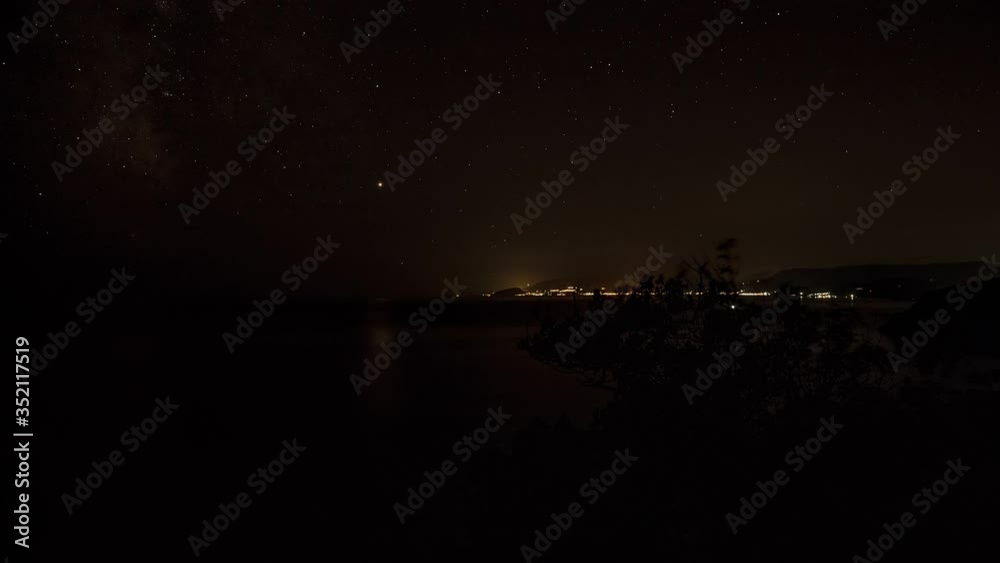 Top view of landscape of bay of mountains over Black Sea with luminous houses and lanterns under bright shining moon and moving starry sky at night in Crimea, time lapse of coastal moonlight path