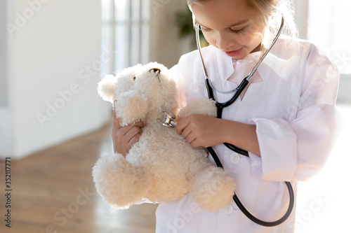 Fotografie Cute small girl kid in whites playing with teddy bear listen heartbeat with stet
