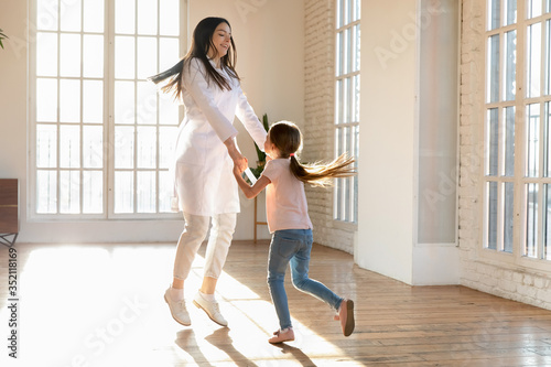 Overjoyed young female nurse and little girl patient have fun dancing in private clinic, happy woman pediatrician or doctor entertain small preschooler child client at consultation in hospital