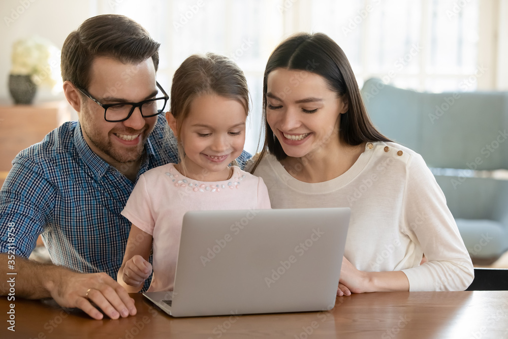 Overjoyed young family with little daughter sit at table watch funny video on laptop together, smiling mom and dad have fun with small girl child using modern computer browsing internet at home Overjoyed young family with little daughter sit at table watch funny video on laptop together, smiling mom and dad have fun with small girl child using modern computer browsing internet at home
