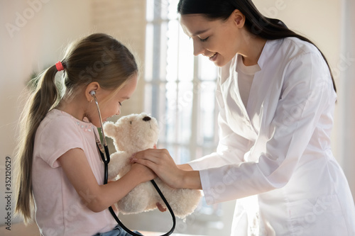 Caring female nurse play with small girl patient, listen to teddy bear heartbeat at regular checkup in clinic, woman pediatrician entertain comfort little child in hospital, healthcare concept