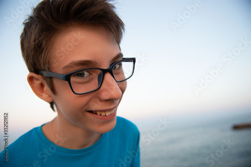 Funny young happy boy laughs looking at the camera  beach at the evening  wide angle. Beautiful smiling spectacled teen boy makes selfie alone at Mediterranean sea coast. Surprise  teenage lifestyle.