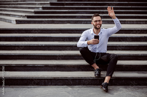 Young cheerful unshaven man elegantly dressed sitting on the stairs outdoors with earphones in ears, holding smart phone and waving to a friend.