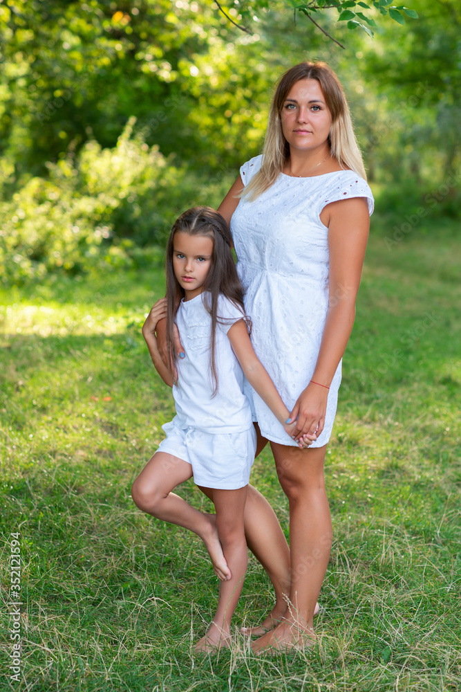 Fototapeta premium Little girl hugs mother, summer nature outdoor. Mom and daughter wearing white clothes against summer greenery. Family walks in park. Trust, kindness, maternity, parenthood, confidence, mother's love