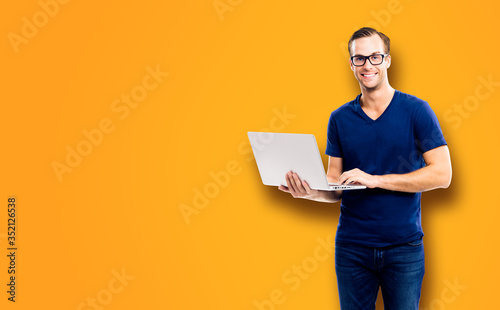 Portrait picture of happy smiling young man in glasses and blue smart casual clothing, holding laptop, standing against loft style wall background.