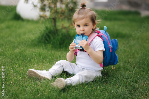 Adorable little girl sitting on the grass with backpack. Cute little girl drinking juise outdoors © Aleksandr