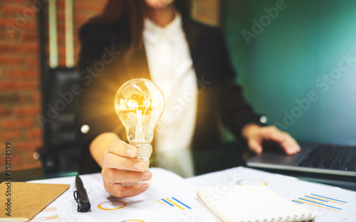 Closeup image of a businesswoman holding a glowing light bulb while working on laptop computer in office