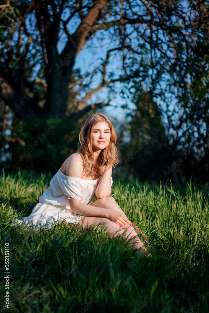 A beautiful young woman is sitting in a field in the green grass. She has a white dress, light hair, bare shoulders, and a happy face. Enjoy nature.