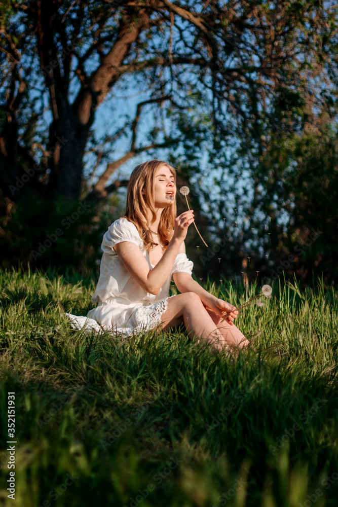 Beautiful Young Woman sitting on the field in green grass and blowing dandelion. Outdoors. Enjoy Nature. Healthy Smiling Girl on summer lawn. Allergy free concept.